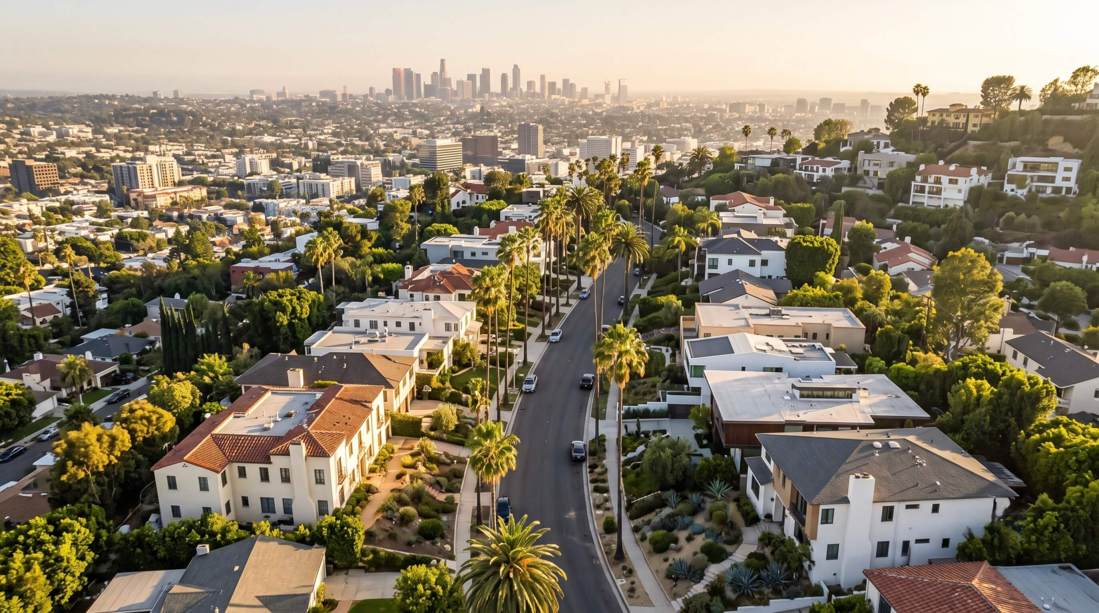 An elevated aerial perspective showcasing diverse residential housing inventory nestled in the Los Angeles hills, emphasizing tree-lined streets in Hollywood Heights with the distant downtown city skyline under golden hour light.